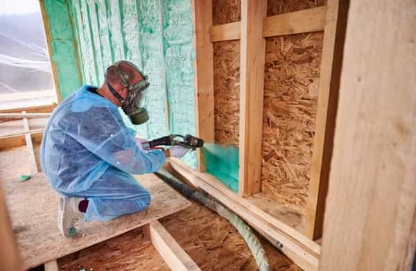 Spray foam technician with mask applying insulation in the wall between wood studs.